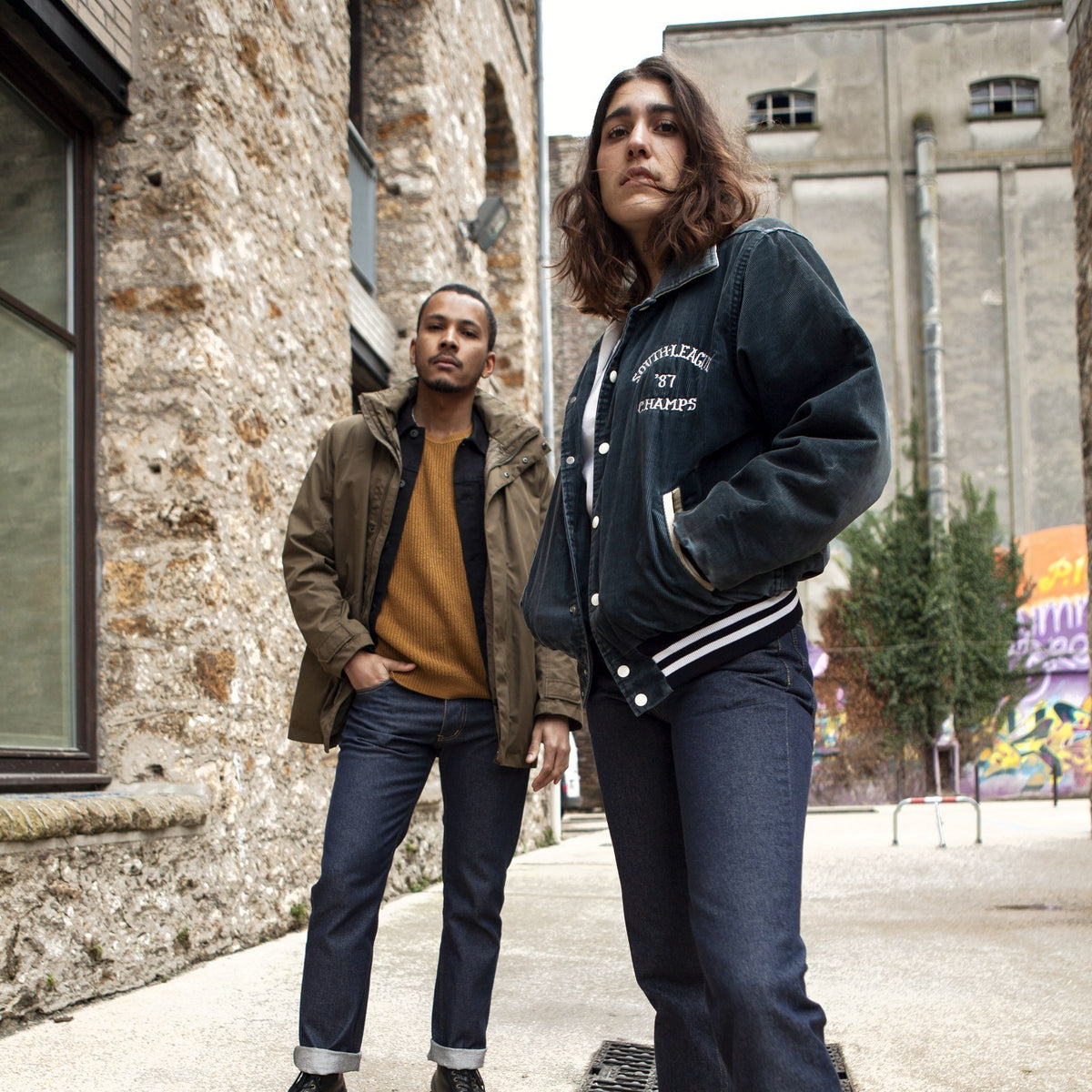 Homme en parka kaki et pull moutarde, femme en teddy noir, tous deux en jean brut Ecclo made in France, dans une ruelle